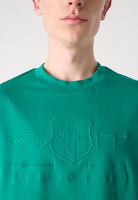 Žalias medvilnės marškinėliai su iškilniu logotipu "GANT" ir "American Sportswear", apvaliu iškirpimu ir trumpomis rankovėmis.