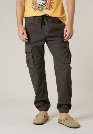 Homme portant un pantalon cargo gris foncé avec des poignets élastiques et des chaussures beige à enfiler, debout devant un fond neutre.