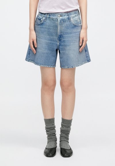 Osoba oblečená v svetlomodrých džínsových šortkách, sivých voľných ponožkách a čiernych baletných balerínkach, stojaca pred jednoliatym bielym pozadím.