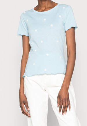 Vrouw draagt een lichtblauw shirt met korte mouwen en kleine witte madeliefjes erop, gecombineerd met witte broek met hoge taille, tegen een effen achtergrond.