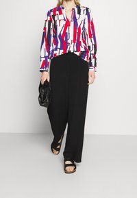 Färgglad blus med mönster som har röda, blå och vita geometriska mönster; matchad med svarta vida byxor och svarta sandaler.