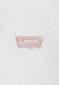 Bijela tkanina s izvezenim ružičastim Levi's logom. Logo se odlikuje karakterističnim zakrivljenim oblikom i urednim vezom na glatkoj teksturi.