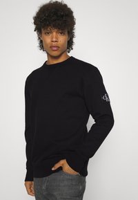 Felpa nera a collo rotondo realizzata in tessuto morbido. Presenta polsini e orlo a coste, con un logo CK applicato sulla manica sinistra.