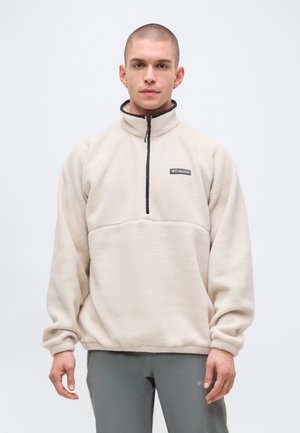 Jeune homme aux cheveux courts portant un pull polaire beige Columbia avec une fermeture éclair noire, debout devant un fond uni.