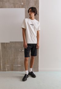 Weißes Baumwoll-T-Shirt mit schwarzem "ellesse"-Logo, kombiniert mit schwarzen Shorts, weißen Socken und schwarzen Schuhen mit gelben Sohlen, in einer minimalistischen Umgebung.