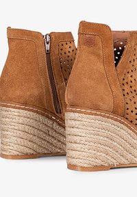 Botas de tobillo de gamuza marrón con cuña, diseño perforado, cremallera lateral y base de paja trenzada. Presenta costuras en color marrón claro y un forro interior suave.