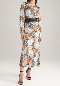 Femme portant une robe maxi à manches longues à motif floral avec une large ceinture noire et des sandales à talons noirs, debout sur un fond neutre.