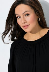 Blusa negra con escote fruncido, que presenta una tela suave y fluida con un acabado texturizado. El cabello cae suelto alrededor de la cara.