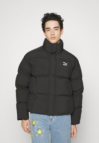 Veste matelassée noire avec col haut, motif matelassé et petit logo blanc de Puma. Le modèle porte un jean bleu clair avec des accents jaunes en forme d'étoile.