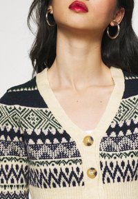 Gestrickte Strickjacke mit V-Ausschnitt, cremefarbener Basis und geometrischen Mustern in Marineblau, Grün und Weiß. Mit zwei großen Knöpfen versehen.