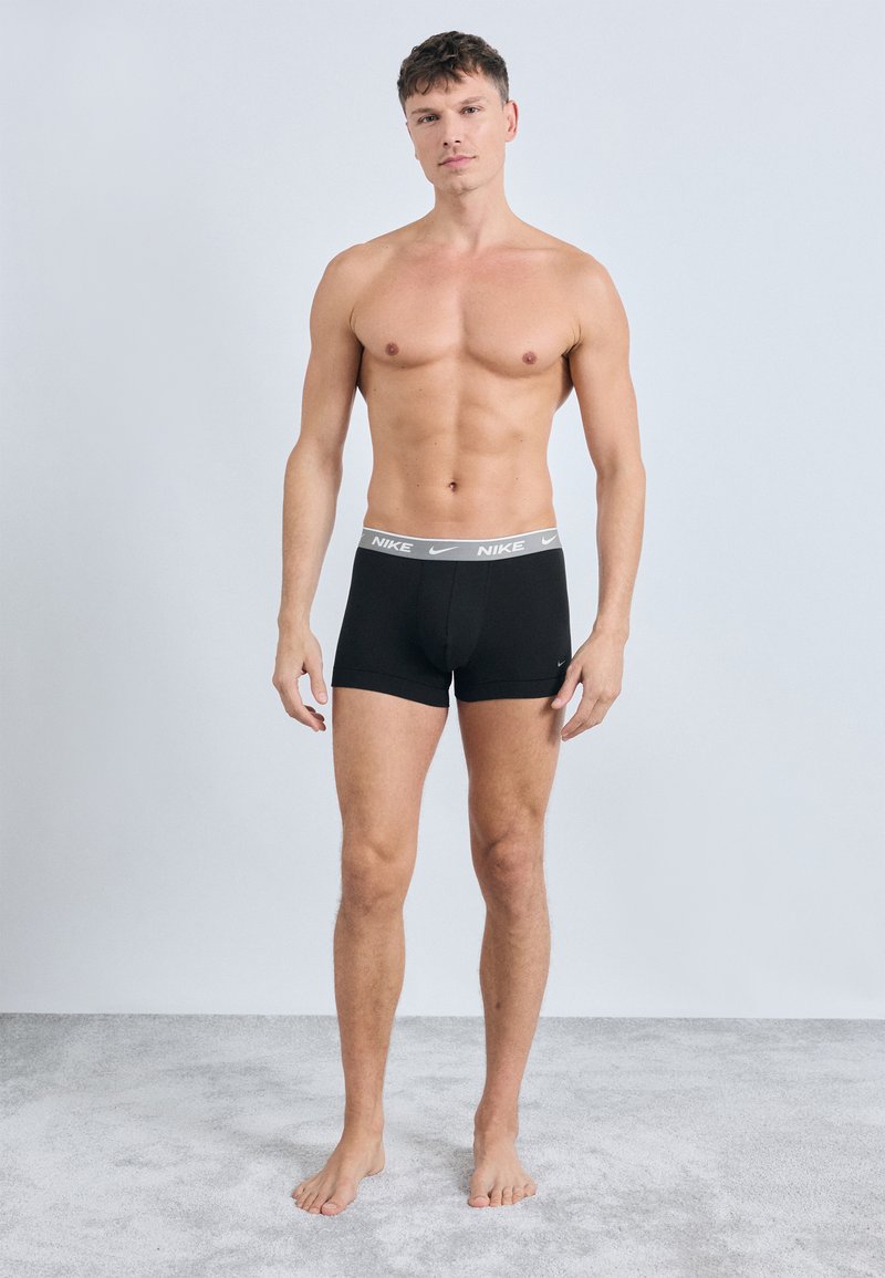 Černé boxerky vyrobené z pružného materiálu, s bílým značkovým pasem. Model stojí naboso na jednobarevném pozadí.
