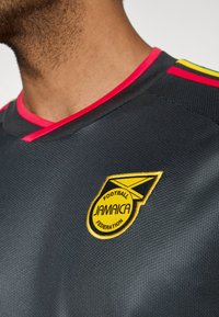 Κοντινό πλάνο ενός ατόμου που φοράει μαύρη ποδοσφαιρική φανέλα με κόκκινο περίγραμμα και κίτρινο λογότυπο της Ομοσπονδίας Ποδοσφαίρου της Τζαμάικας στο στήθος.