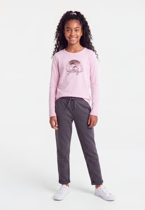 Fille aux longs cheveux bouclés attachés en arrière, souriante, debout, portant un t-shirt rose à manches longues avec un motif, un pantalon gris foncé et des baskets blanches.