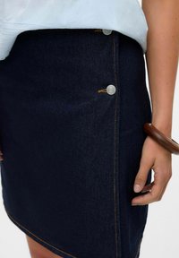 Denimkjol i mörkblått med en figursydd design. Har silverfärgade knappar och kontrasterande guldstickningar längs sömmarna.