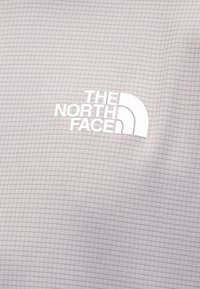 Λευκό λογότυπο "The North Face" σε ανοιχτό γκρι ύφασμα με μικρό σχέδιο πλέγματος, πιθανώς μέρος εξωτερικού ρουχισμού.