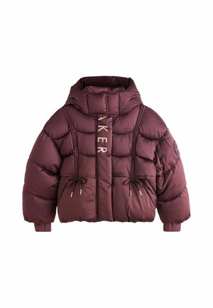 Junge Person mit lockigem Haar, die eine kastanienrote Steppjacke, lockere blaue Jeans und weiße Turnschuhe trägt und auf einem grauen Boden steht.