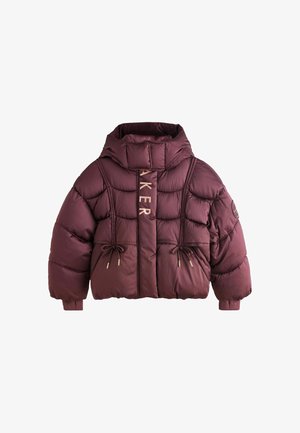 Junge Person mit lockigem Haar, die eine kastanienrote Steppjacke, lockere blaue Jeans und weiße Turnschuhe trägt und auf einem grauen Boden steht.