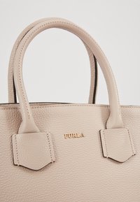 Beżowa skórzana torebka z dwoma teksturowanymi uchwytami i złotym brandingiem. Charakteryzuje się prostym wzornictwem oraz widocznymi akcentami szwów.