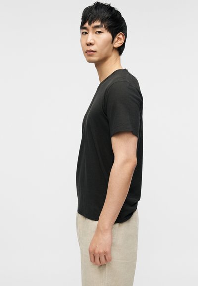 Σκούρο γκρι κοντομάνικη μπλούζα με στρογγυλή λαιμόκοψη, εμφανιζόμενη σε ανδρικό μοντέλο που στέκεται πλαγίως μπροστά σε απλό φόντο.