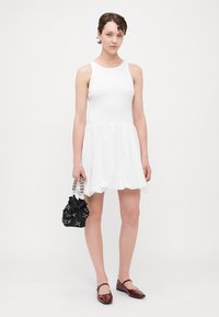 Weißes ärmelloses Kleid mit enganliegendem Oberteil und weit ausgestellt, strukturiertem Rock. Schwarze Bucket Bag mit dekorativen Akzenten. Dunkelrote Ballerinas.