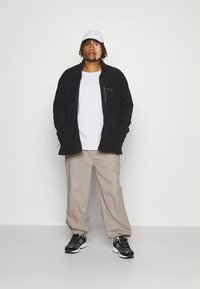 Czarna bluza polarowa z zamkiem błyskawicznym, jasnoszary t-shirt, beżowe spodnie oraz czarne trampki. Biała czapka typu baseball. Codzienny strój, luźny krój.