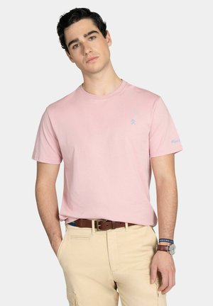 Joven con camiseta rosa claro, pantalones beige, cinturón marrón y reloj de pulsera, con un fondo claro neutro.