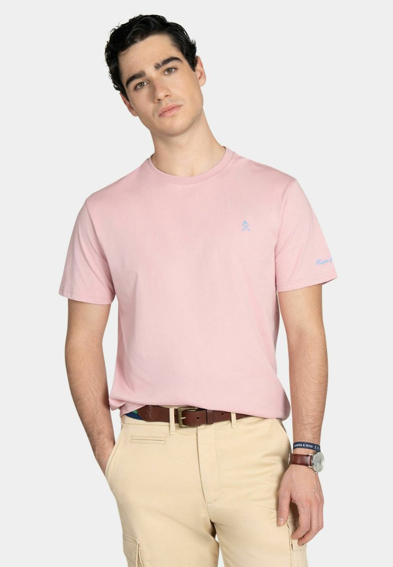 Jeune homme portant un t-shirt rose pâle, un pantalon beige, une ceinture marron et une montre-bracelet sur un fond clair neutre.