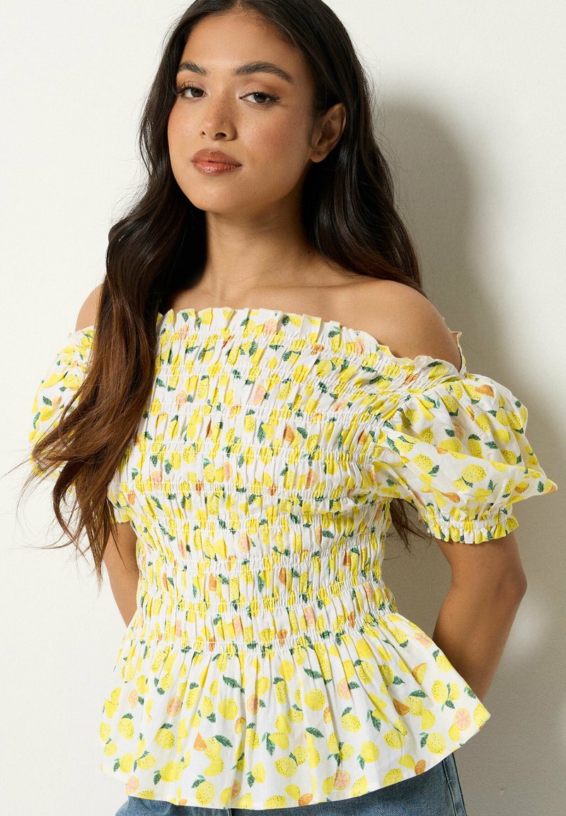 Mujer joven con cabello largo y oscuro, vistiendo un top blanco sin hombros con estampado de limones amarillos y mangas abullonadas, posando frente a un fondo liso.