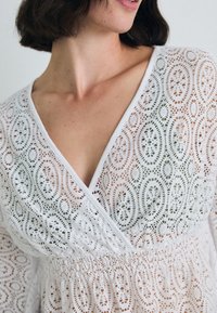 White lace dress featuring an intricate oval pattern, deep V-neckline, and gathered waist detailing, made from lightweight, breathable fabric.