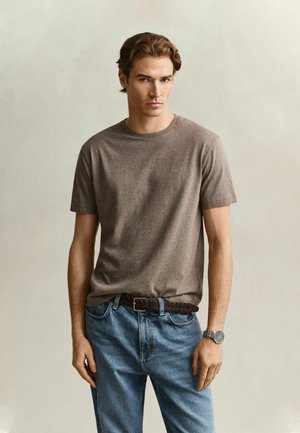 Jeune homme aux cheveux châtain clair portant un t-shirt marron à manches courtes, un jean bleu, une ceinture tressée foncée et une montre-bracelet argentée, debout devant un fond uni.