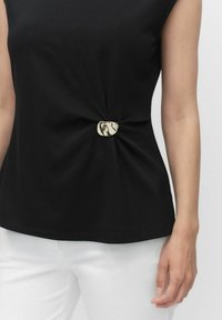 Zwarte T-shirt met een twistdetail aan de voorkant, voorzien van een glanzende metallic accent. G gedragen met witte broek, met een gladde, zachte textuur.