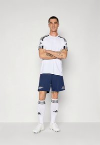 Camiseta deportiva blanca con rayas negras en las mangas, combinada con pantalones cortos azul marino que llevan un logo. El conjunto se completa con calcetines blancos y tacos.