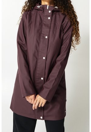 Femme portant un long imperméable violet foncé avec des boutons-pression argentés et une capuche, debout les mains jointes devant.