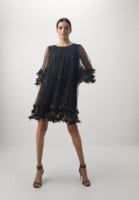 Nainen seisoo mustassa läpinäkyvässä mekossa, jossa on kukkakoristeet hihoissa ja helmassa, jalassaan mustat korkokengät, taustalla on yksinkertainen tausta.