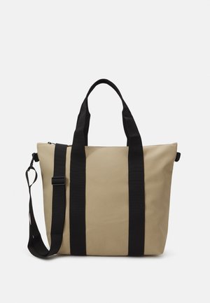 Sac fourre-tout beige fabriqué en tissu résistant avec des poignées en sangle noire, doté d'une fermeture éclair, d'une forme structurée et d'un design minimaliste.