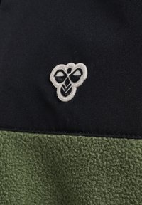 Hvid og sort broderet bi-logo på et sort stofpanel over et tekstureret olivengrønt stofafsnit.
