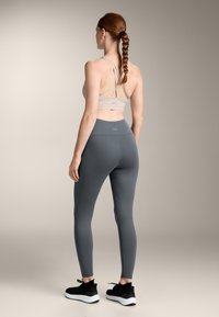 Svetlo siva športna modrčka s križnimi naramnicami, v kombinaciji s temno sivo leggingi in črnimi teniskami, z gladkim in elastičnim materialom.