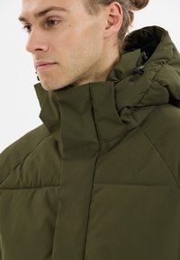 Olijfgroen pufferjack met een hoge kraag, voorzien van een gestikte uitstraling en verstelbare trekkoorden aan de kap en nek voor een stevige pasvorm.
