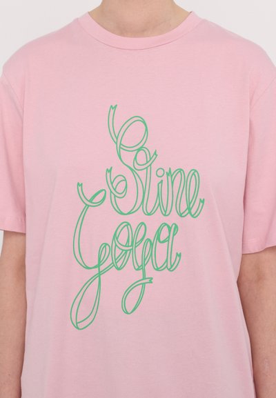 T-shirt de algodão rosa com texto em verde cursivo "Sine Yoga" na parte da frente. Gola redonda e mangas curtas. Design simples e casual.