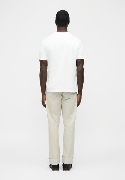 Άνδρας που στέκεται με την πλάτη γυρισμένη, φορώντας απλό λευκό κοντομάνικο μπλουζάκι, μπεζ παντελόνι με γυρισμένα ρεβέρ και μαύρα παπούτσια σε λευκό φόντο.