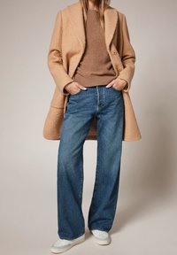 Beige textuurjas, bruine trui en wijdvallende blauwe spijkerbroeken. Witte sneakers maken de outfit af, met een ontspannen pasvorm en casual stijl.