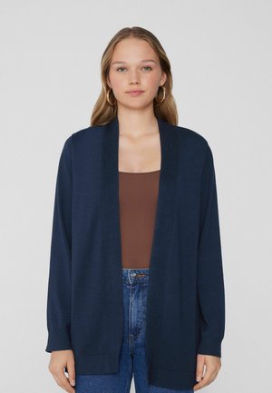 Námořnický cardigan vyrobený z měkké tkaniny, s otevřeným předním designem, dlouhými rukávy a pružnými akcenty, kombinovaný s hnědým topem a modrými džíny.