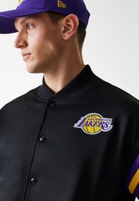 Black varsity jacket with a Los Angeles Lakers logo. Features a ribbed collar, buttons, and purple accents on the sleeves.
