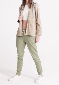 Khaki kurtka z szarymi akcentami, biała prążkowana bluzka do brzuchem i zielone dresy. Model nosi białe sneakersy; luźny krój, lekki materiał.