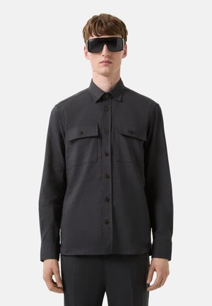 Dark grey, long-sleeve shirt with a button front, two chest pockets, and brown buttons. Smooth fabric with a structured fit.