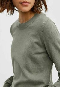 Pull vert clair avec une finition texturée, présentant un col festonné et une coupe ajustée. Tissu tricoté doux, idéal pour être superposé.