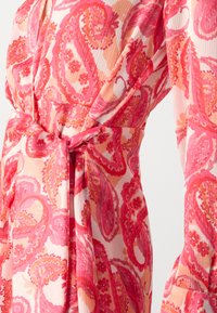 Robe longue avec un motif paisley rose sur un fond clair, présentant une texture plissée et un détail de taille nouée.