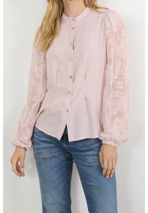 Woman wearing a light pink buttoned blouse with sheer lace floral sleeves and blue jeans, standing against a plain white background.