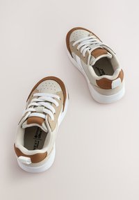 Baskets beige et tan avec lacets blancs, design à perforations sur la pointe, et accents marron contrastants sur le talon et les panneaux latéraux. Semelle blanche.