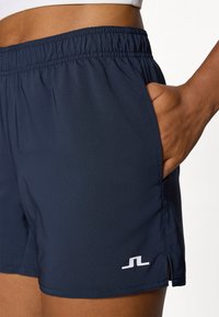 Calções atléticos azuis-marinho com uma cintura elástica, bolsos laterais e detalhe de logótipo branco. Apresenta um tecido leve e respirável.